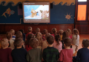 Na zdjęciu dzieci oglądające spektakl on line wyświetlany na tablicy multimedialnej